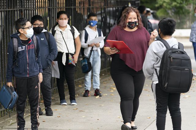 A masked woman carrying a red clipboard speaks to a student outside PS 179 in Kensington, as several students lined up and wearing masks and backpacks look on.
